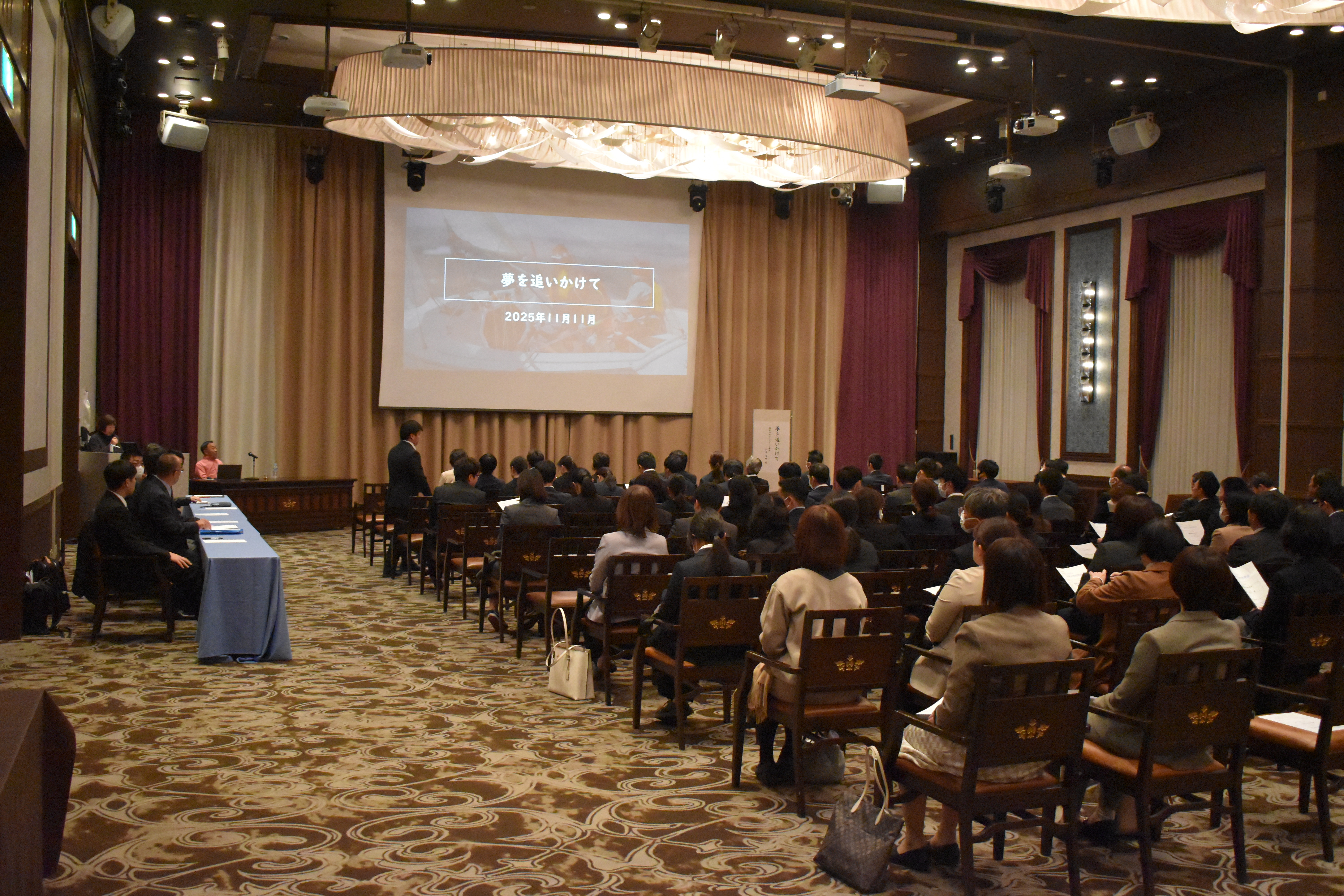 写真：講演会場で大きなスクリーンにタイトルが映され、参加者が発表を聞いている様子