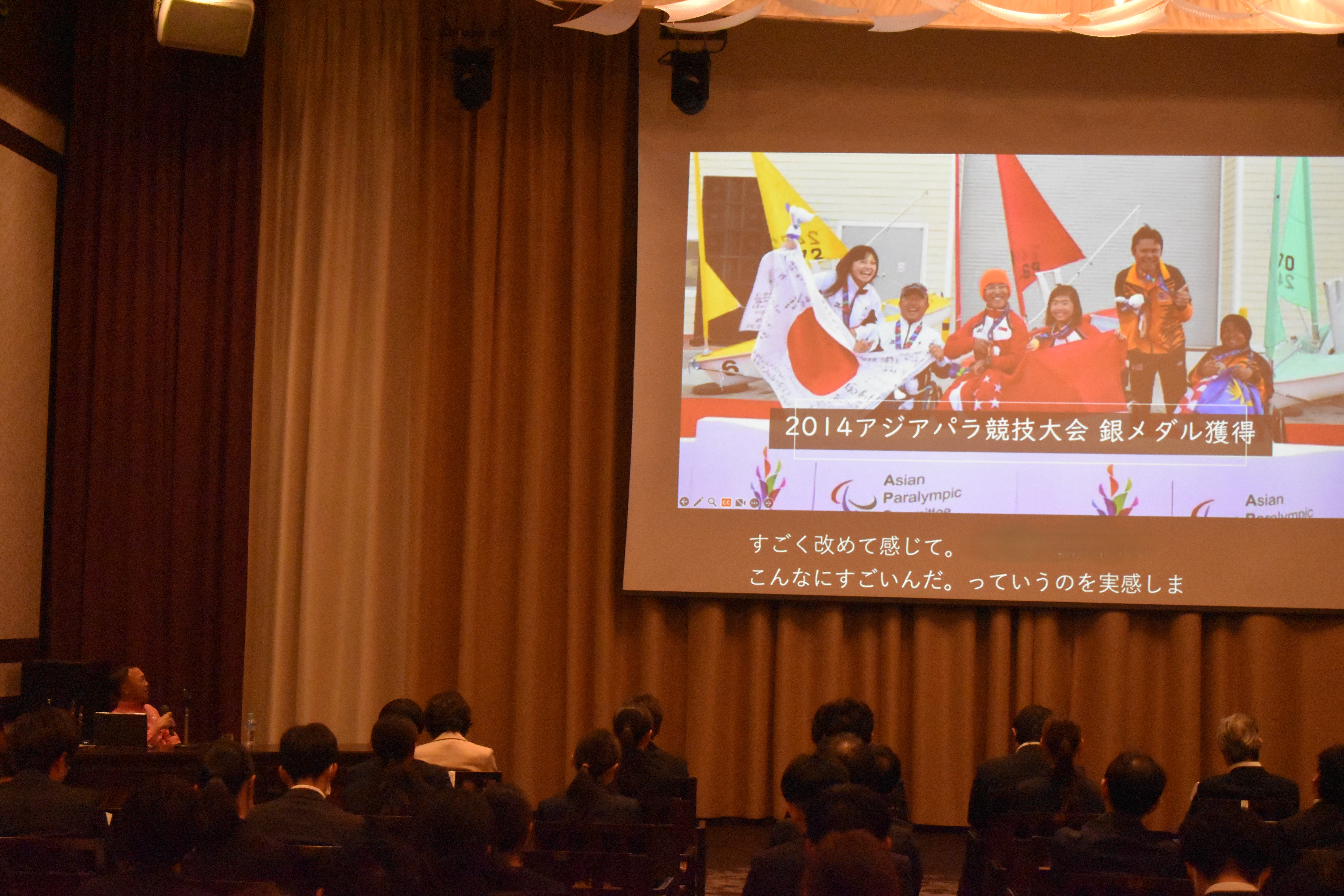 写真：スクリーンに大会の表彰シーンが映され、参加者が注目している講演の様子