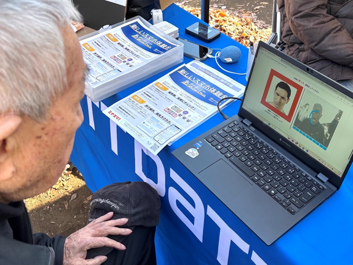 写真：屋外イベントで高齢者がノートPCの画面を見て、横にスマホ安全利用の資料が並ぶ